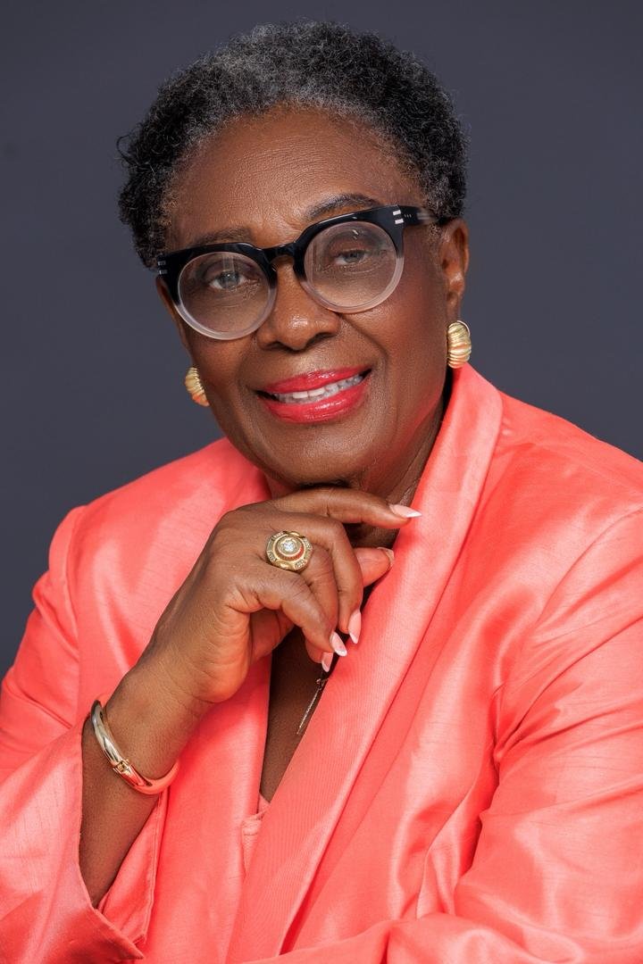 Close portrait of Chief Mrs. Josephine Chinyelu Nwosu wearing glasses and a coral jacket.